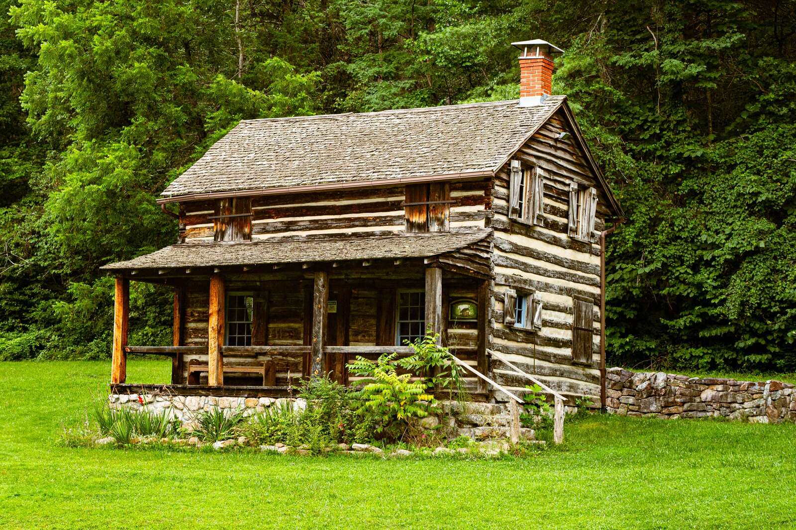 Log cabin with a stone wall below a wooded hillside